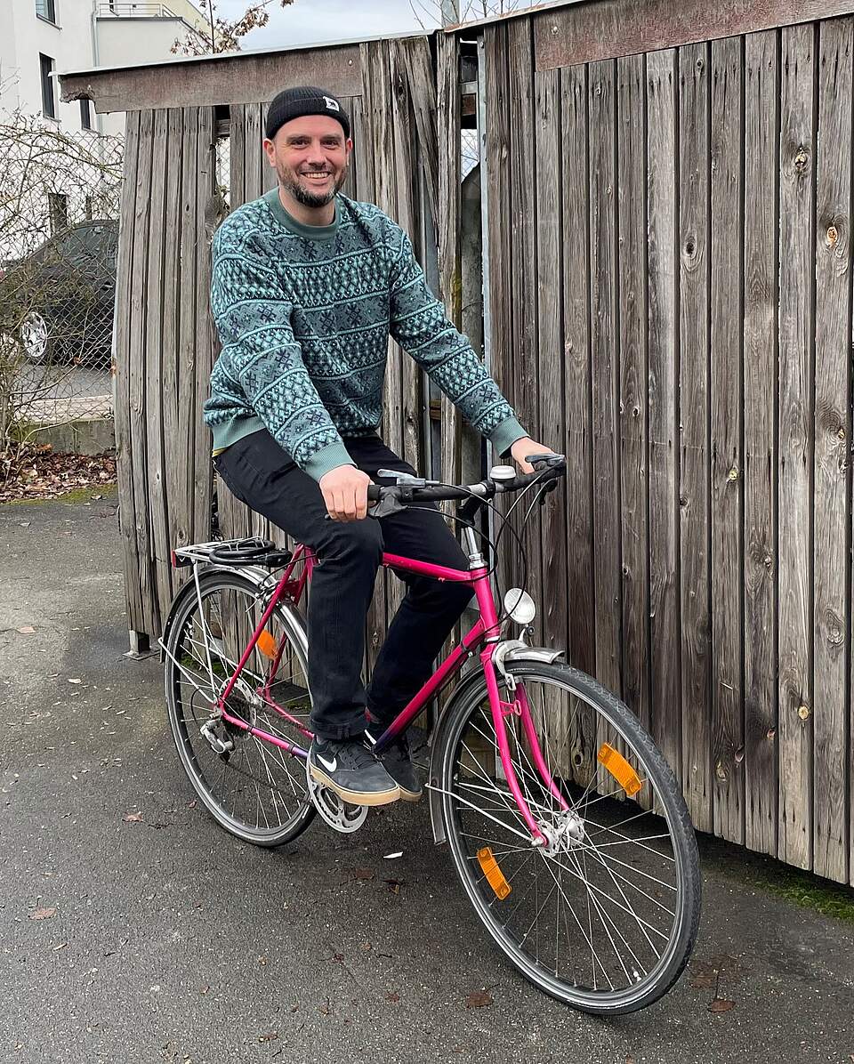Forchheims neuer Radverkehrsbeauftragte der Stadt: Tobias Wilhelm Radverkehrsbeauftragter der Stadt Forchheim Tobias Wilhelm sitzt auf einem pinkfarbenen Herrenrad vor einer hölzernen Bretterwand.