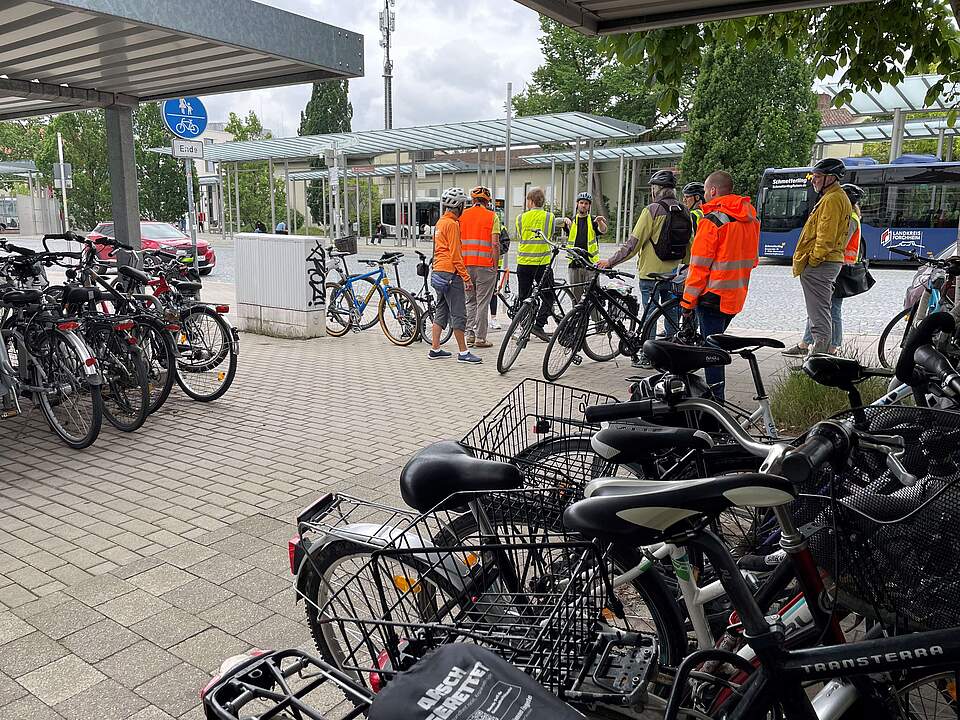 Bewertungskommission der AGFK Bayern und Vertreter der Stadt Forchheim bei der Bestandserfassung der Radverkehrsförderung. Radfahrergruppe mit Helmen sowie geleben und orangefarbenen Warnwesten stehen mit ihren Rädern an einem Fahrradabstellplatz vor einem Busbahnhof.