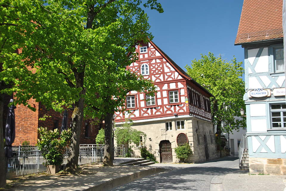 Eine mit Kopfsteinpflaster ausgelegte Straße in einer Altstadt führt durch historische Fachwerkhäuser. Links säumt eine Baumallee die Straße.