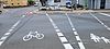 Weiße Straßenmarkierungen zeigen einen Fußgängerüberweg und einen in eine Straße einmündenden Fahrradweg mit einer roten Warnmarkierung an der Einmündung.