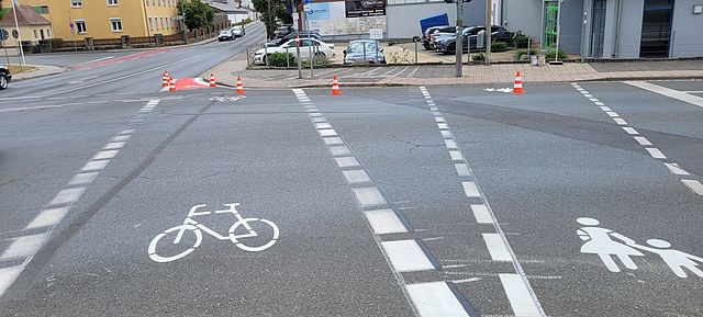 Weiße Straßenmarkierungen zeigen einen Fußgängerüberweg und einen in eine Straße einmündenden Fahrradweg mit einer roten Warnmarkierung an der Einmündung.