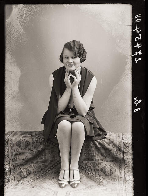 Eine schwarz-weiß Fotografie aus den 1920er Jahren zeigt eine Frau sitzend und mit auf Händen aufgestützten Kinn, die lächelnd in die Kamera blickt.