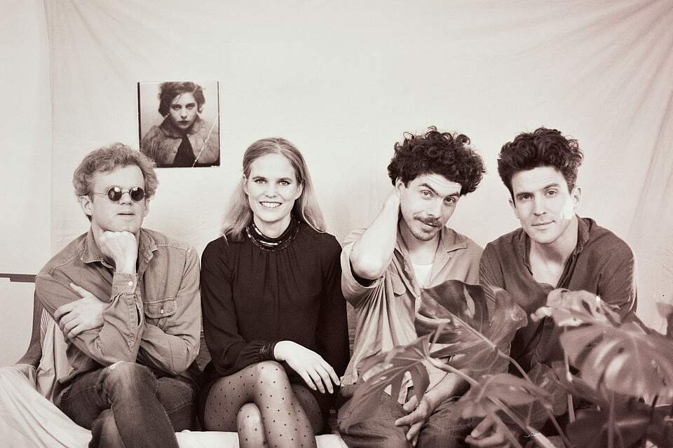 Die Band DOTA, die im KulturSommerQuartier auftritt. Die Künstlerin Dota Kehr umrahmt von ihren drei Bandmitgliedern. im Hintergrund hängt ein älteres Portraitfoto einer Dame, rechts im Vordergrund ragen die Blätter einer Zimmerpflanze in das Foto.