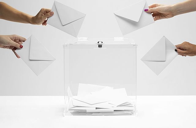 Eine transparente Wahlurne steht auf einem weißen Tisch. Vier Hände strecken Briefumschläge in Richtung der Wahlurne.