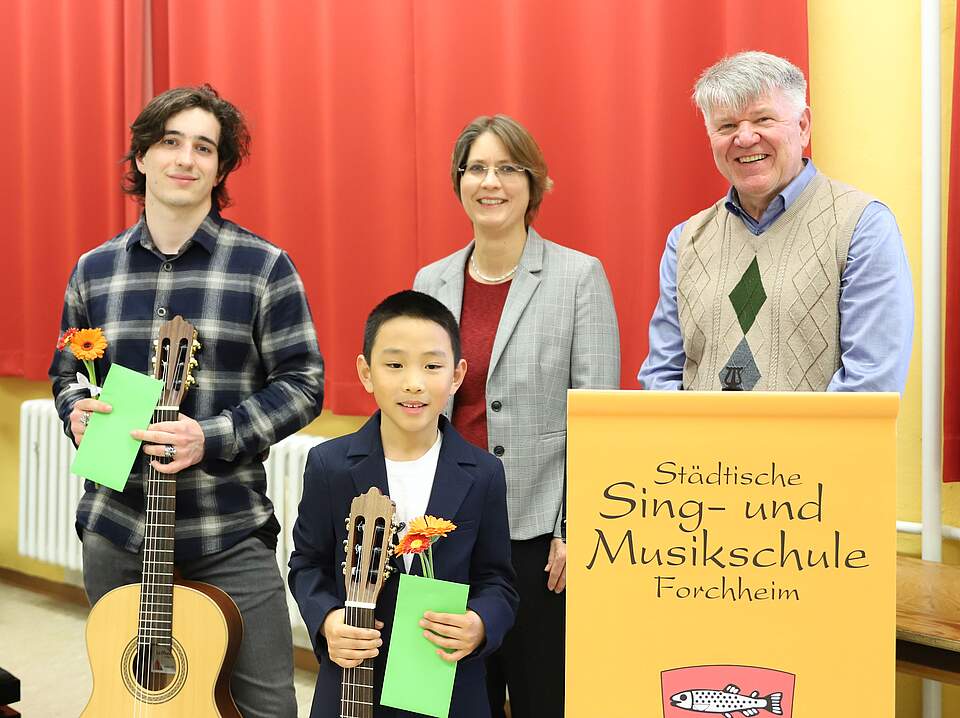 Preisträger des Regionalwettbewerbs "Jugend musiziert" 2024 Die beiden Gewinner des Wettbewerbs mit Bürgermeisterin Dr. Annette Prechtel und dem Leiter der der Städtischen Sing- und Musikschule, Jürgen Hiltl vor einem roten Theatervorhang. Rechts im Bild steht ein auf einem gelben Plakat Städtische Sing- und Musikschule und ein Teil des städtischen roten Wappens mit Forellen.