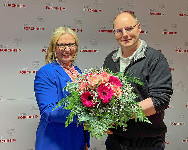 Die neue Forchheimer Bürgermeisterin Martina Hebendanz steht mit einem Blumenstrauß zusammen mit Wahlleiter Andreas Schneider vor einer Pressewand mit dem Logo der Stadt Forchheim.