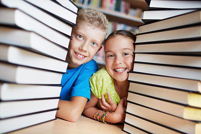 Ein Junge und ein Mädchen schauen zwischen zwei Buchstapeln hervor. Im Hintergrund befinden sich ein Bücherregal.