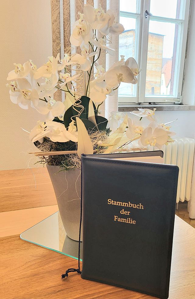 Ein Familienstammbuch steht aufgestellt auf einem Tisch neben einem weißen Blumenschmuck.