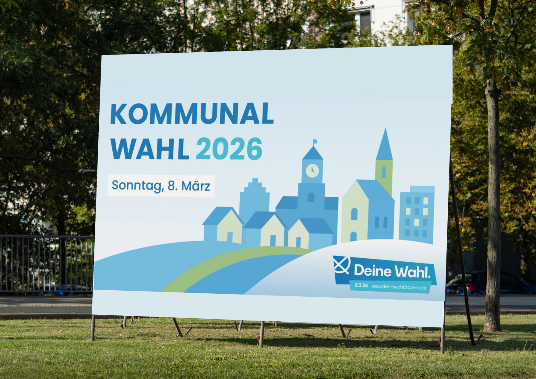 Ein Großplakat auf einer Grünfläche an einer Straße zeigt eine Grafik mit einer Ortschaft und einer Hügelkette. Links oben im Bild steht: "Kommunalwahl 2026, Sonntag, 8. März". Rechts unten im Bild sieht man den Schriftzug mit einem Kreuz: "Deine Wahl - 8.3.26 -www.deinewahl.bayern.de