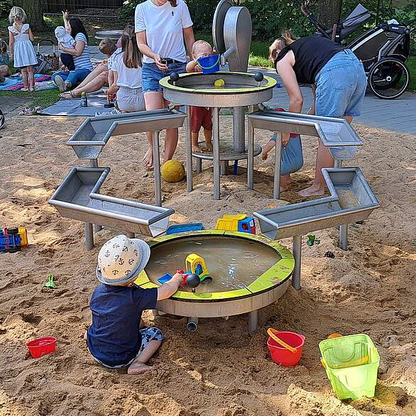 Mütter mit ihren Kindern an einem Sandspielplatz mit Wasserspielanlage.
