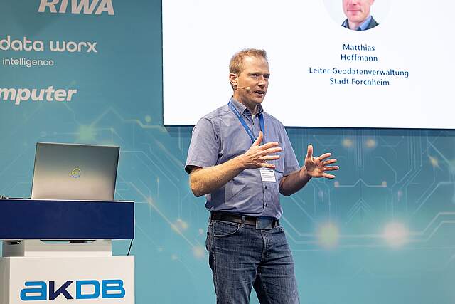 Matthias Hoffmann von der Geodatenverwaltung der Stadt Forchheim hält einen Vortrag zum Digitalen Zwilling auf der Messe Smart Country Convention 2025 in Berlin Ein Mann spricht auf einer Bühne zu einem Publikum. Links neben ihn befindet sich ein Rednerpult im Hintergrund das Logo der Firma RIWA auf einem blauen Hintergrund.