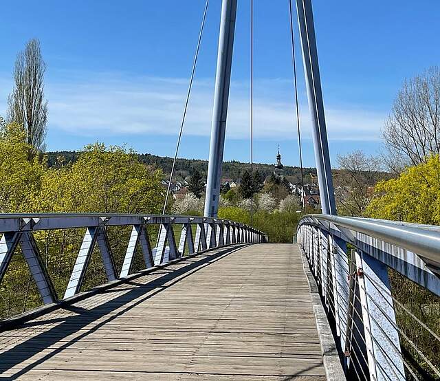 Eine Fußgängerbrücke aus Stahl und Holz. Am Ende dr Brücke befinden sich große Bäume. Im Hintergrund sieht man mehrere Häuser und einen Kirchturm in einiger Entfernung.