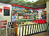 Ein historischer Kaufladen im Stil der 50er Jahre mit Produkten und Marken aus dieser Zeit in den Regalen. Im Vordergrund steht eine gläserne Theke mit einer manuellen Lebensmittelwaage und verschiedene Kuchen, Likörflaschen und Keksdosen.