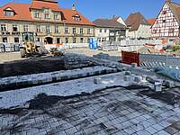Ein großer Platz umgeben von Häusern wird neu gepflastert. Es sind Absperrungen, Baumaterial und ein Bagger zu sehen.