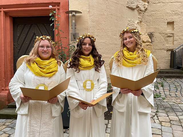 Drei junge Frauen in weißen Engelskostümen und goldenen Haarkränzen halten jeweils ein goldenes offenes Buch in ihren Händen.