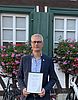Oberbürgermeister Dr. Uwe Kirschstein hält eine Urkunde in den Händen. Im Hintergrund steht ein Fachwerkgebäude mit buntem Sommerblumenschmuck.
