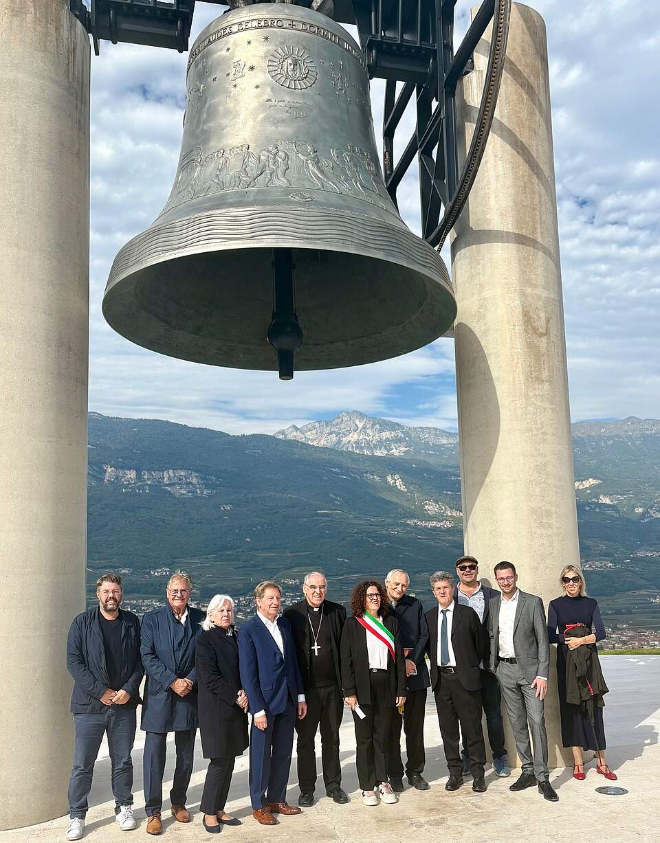 100. Jubiläum der Friedensglocke in Rovereto - Delegation mit Vertretern Roveretos unter der Friedensglocke Die Forchheimer Delegation zusammen mit Vertretern der Stadt Rovereto und der Kirche unter der Friedensglocke. Im Hintergrund Berge und blauer Himmel mit Wolken.