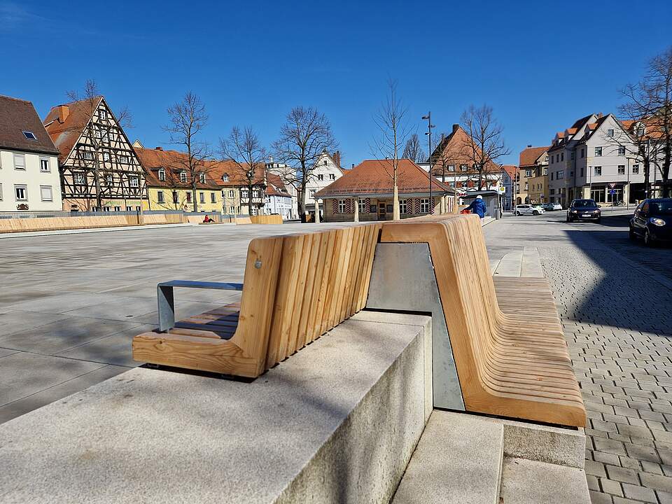 Ein großer, moderner Stadtplatz mit Sitzmöbel aus Holz. Im Hintergrund das historische, einstöckige Sandtsteingebäude "Alte Wache". Die Platz ist von Häusern umgeben.