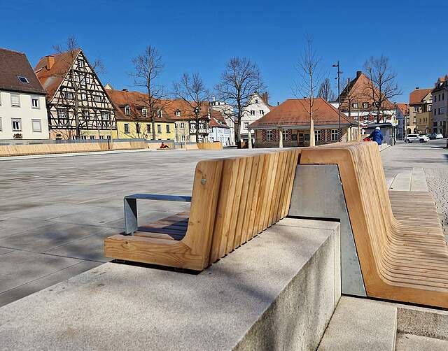 Ein großer, moderner Stadtplatz mit Sitzmöbel aus Holz. Im Hintergrund das historische, einstöckige Sandtsteingebäude "Alte Wache". Die Platz ist von Häusern umgeben.