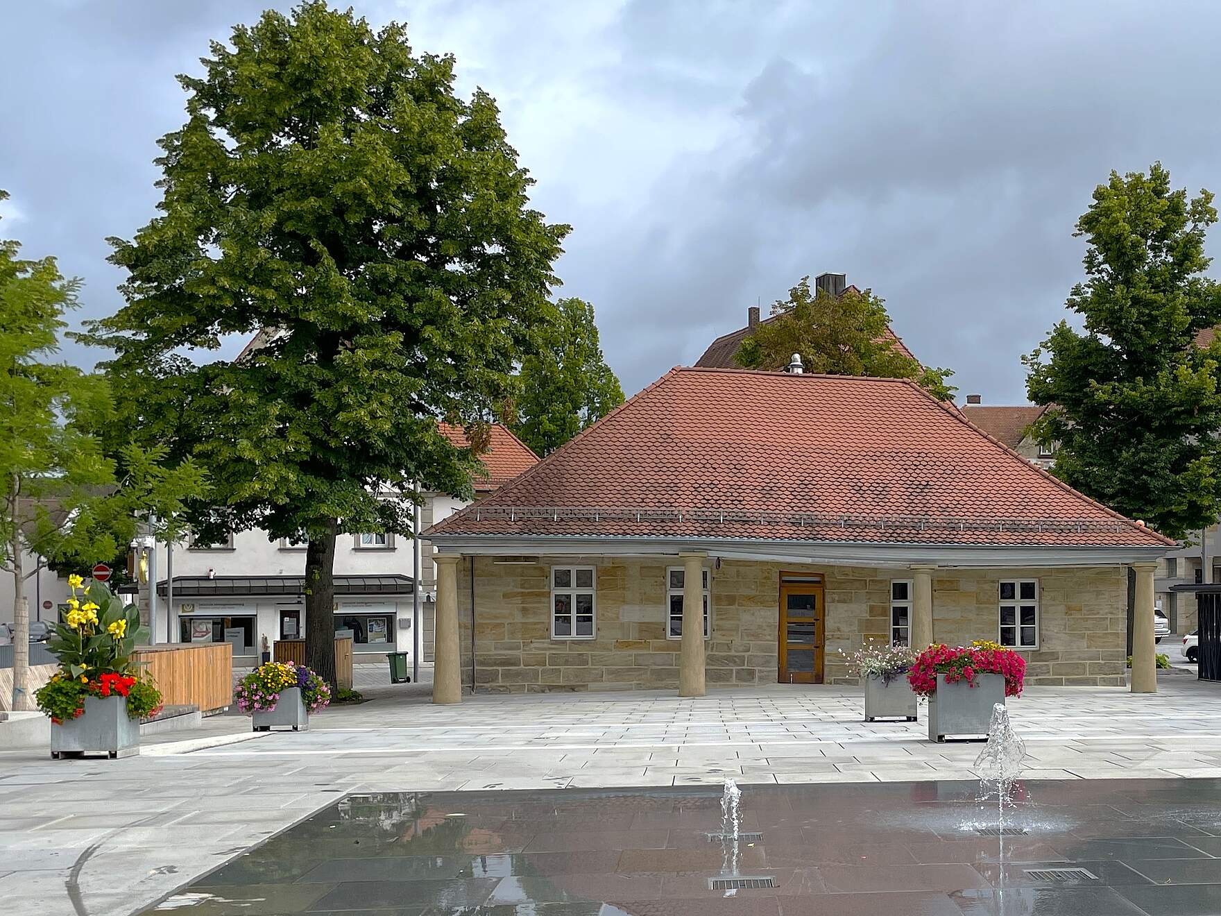 Ein einstöckiges Sandteingebäude mit vier Steinsäulen auf einem Stadtplatz umgeben von Bäumen, Wasserspiel und bunten Pflanzkübeln.
