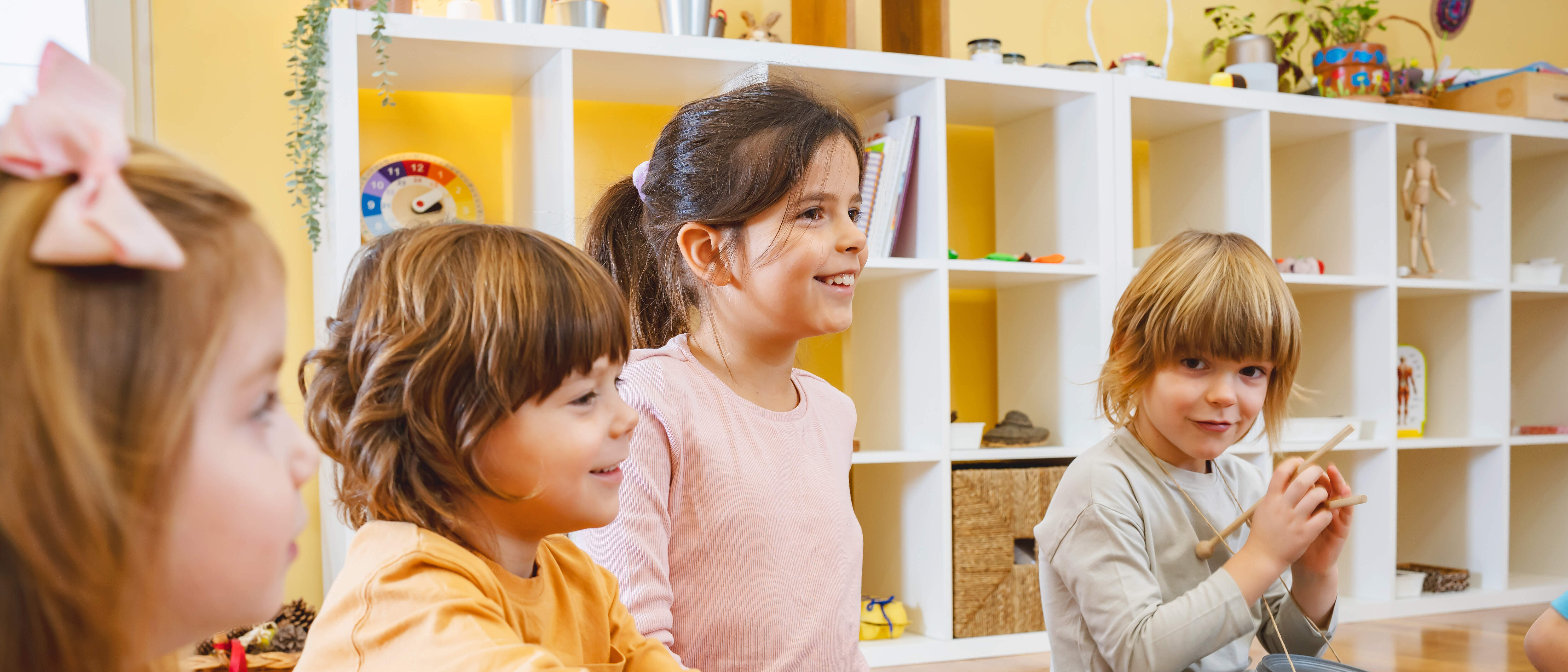Vier Kindergartenkinder mit verschiednenen Musikinstrumenten sitzen nebeneinander auf dem Boden eines Kindergartenraumes. Im Hintergrund befindet sich ein weißes Regal mit Spielsachen.