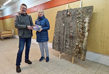 Ein Mann und eine Frau stehen links neben einem sogenannten Sedimenttransferpräparat, das an einer Wand lehnt. Das Präparat zeigt anhand von Gesteinsschichten anschaulich Forchheims Geschichte.