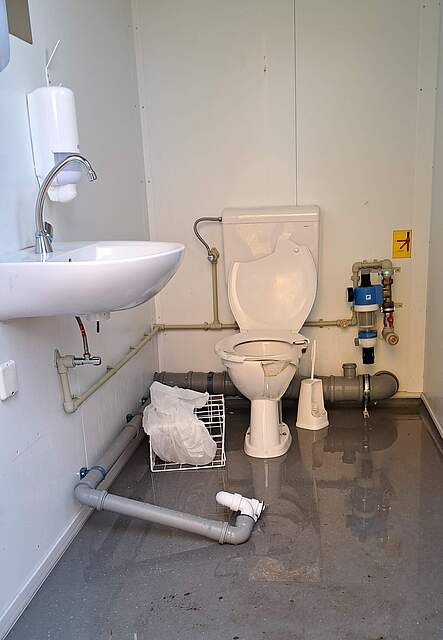 Eine zerstörte Toilettenanlage in einem Container. Bruchstücke der Toilettenschlüssel und ein abgerissenes Wasserrohr liegen über den Boden verteilt.