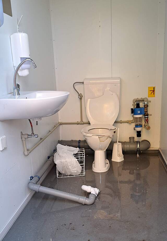 Eine zerstörte Toilettenanlage in einem Container. Bruchstücke der Toilettenschlüssel und ein abgerissenes Wasserrohr liegen über den Boden verteilt.