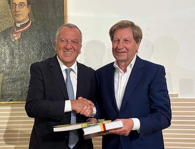 Partnerschaftsbeauftragter Reinhold Otzelberger bei der Übergabe des Buches „Wehrhaftes Forchheim“ mit persönlicher Widmung an den Vorsitzenden Marco Marsilli, Vorsitzender der Stiftung Campana dei Caduti. Im Hintergrund an der Wand ein historisches Portrait.