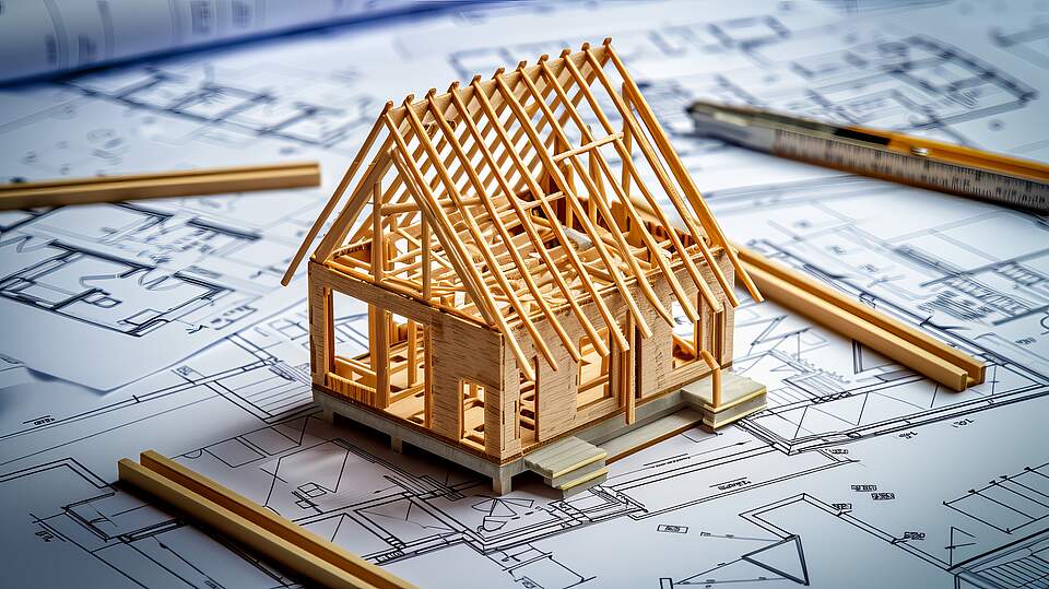 Ein kleines Modell eines Hausrohbaus aus Holz steht auf mehreren Bauplänen. Um das Modell herum liegen noch schmale Holzleisten und links oben schemenhaft einen Zollstock.