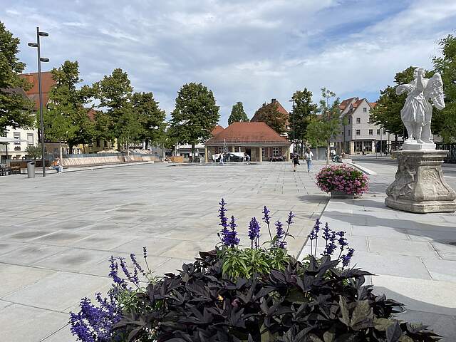 Paradeplatz Forchheim Ansicht des umgestalteten Paradeplatzes mit neun Bäumen, bunt bepflanzten Blumenkübeln, Stadtmöbeln, einem Wasserspiel und der Mariengruppe (Statuen). Am Platzende befindet sich die sogenannte "Alte Wache", ein historisches Sandteingebäude mit Eingangssäulen.