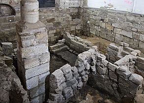 Man sieht archäologische Ausgrabungen in einer großen historischen Halle mit Mauern und einer Säule.