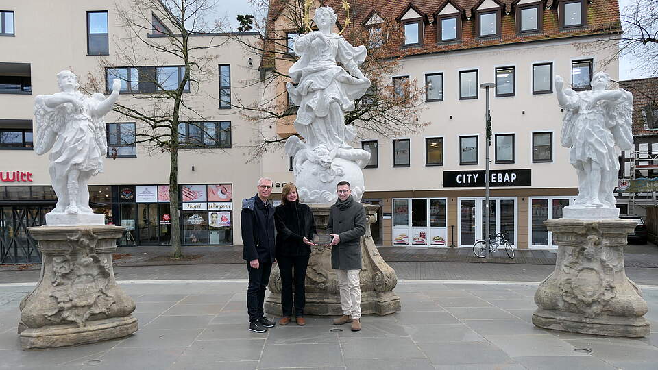Mariengruppe am Paadeplatz Drei Personen stehen bei einer historischen Figurengruppe auf einem Stadtplatz und präsentieren ein Messingschild mit der Aufschrift "Heimatverein Forchheim - Restauriert 2024 - gefördert vom Heimatverein Forchheim Ofr. e.V." .