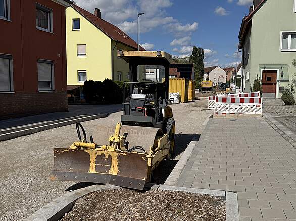 Neugestaltung Bammersdorfer Straße Bauabschnitt 1 mit Kreuzung Von-Ketteler-Straße Baufahrzeuge, Container und Absperrbarken an einer Straßenbaustelle in der Bammersdorfer Straße nahe dem Kreuzungsbereich der Von-Ketteler-Straße. Der geteerte Fahrbahnbelag fehlt noch, der Untergrund ist bereits geschottert und verdichtet.