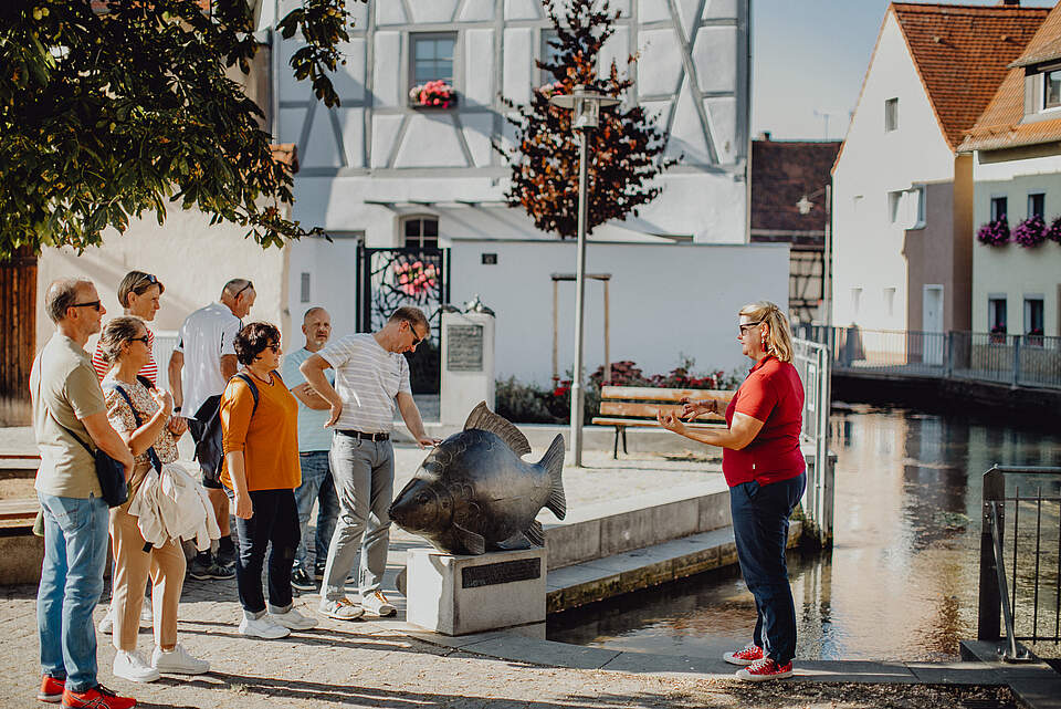 Eine Gästeführerin erläutert einer Gruppe Touristen ein städtisches Wahrzeichen "Fisch" an der Wiesent an einem sonnigen Tag.