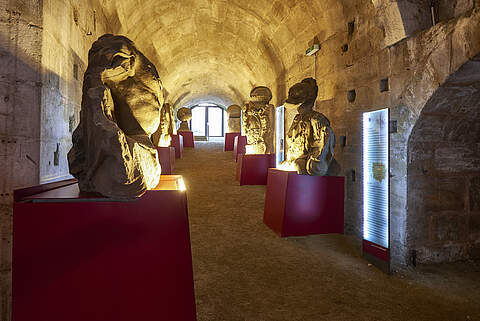 Erlebnismuseum Rote Mauer Mehrere Steinbüsten auf roten Erhöhungen befinen sich an den Seiten eines tunnelartigen Gewölbes. Sanftes Licht erhellt den Gang mit beleuchteten Infotafeln.