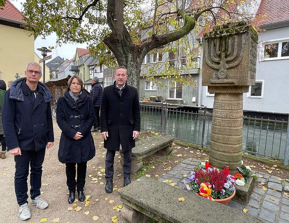 Reichpogromnacht 2024: Die Forchheimer Bürgermeister gedenken an die jüdischen Mitbürgerinnen und Mitbürger. Zwei Männer und eine Frau in dinkler Kleidung stehen an einem Synagogendenkmal. Eine bunt bepflanzte Blumenschale steht am Fuße des Denkmals. Im Hintergrund befinden sich ein grpßer Baum und Häuser.