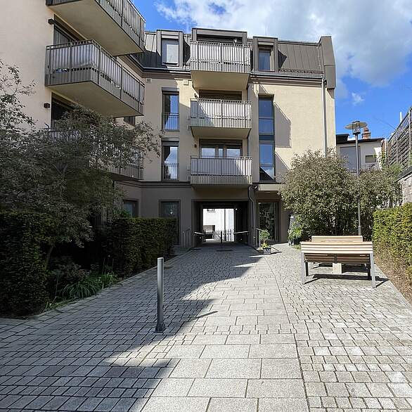 Gepflasterter Eingangsbereich eines dreistöckigen Wohnkomplexes mit STräuchern und Büschen sowie einer Tisch-/Bankgruppe zum Verweilen.