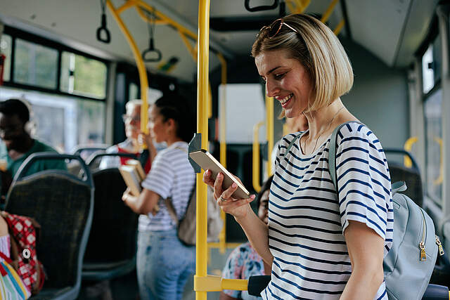 Menschen in einem Bus sitzend und stehend. Im Vordergrund steht eine blonde Frau und schaut auf ihr Smartphone.
