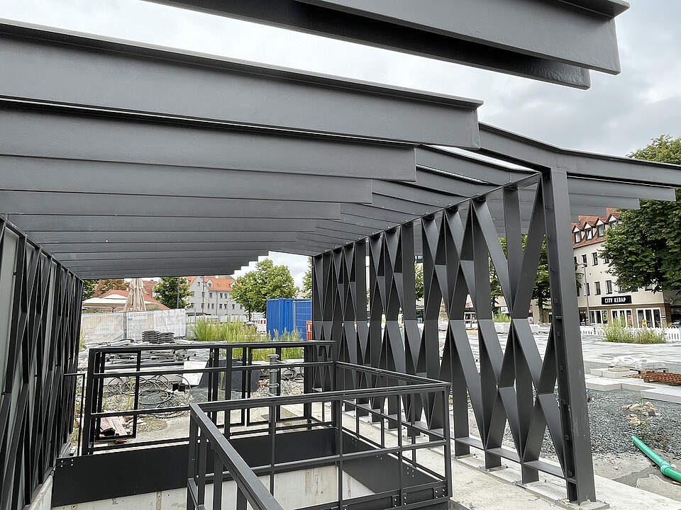 Ein moderner Hochbau aus dunklem Stahl an einem Treppenabgang zu einer Tiefgarage errichtet. Umliegend befindet sich eine Großbaustelle mit Absperrungen und verschiedenem Baumaterial.