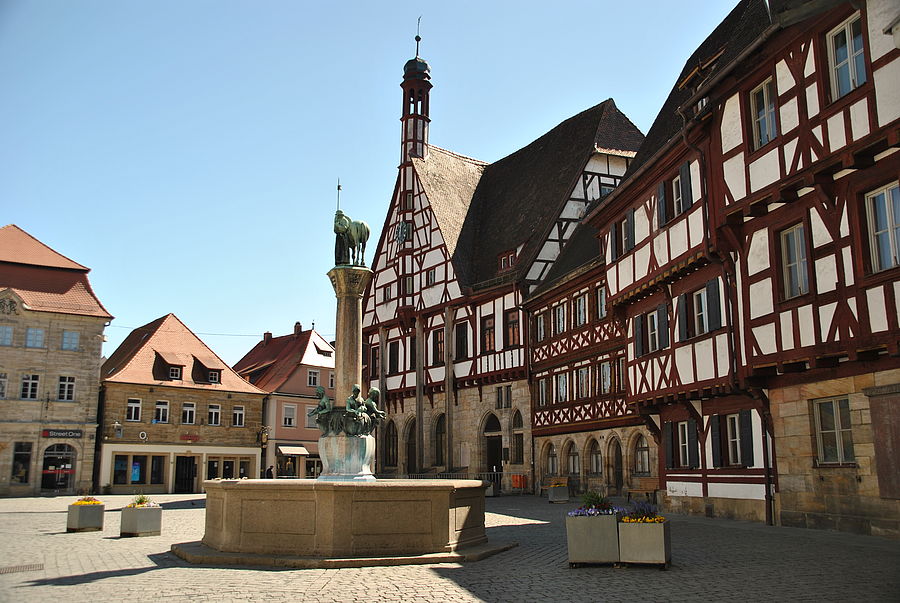 zu sehen sind das Fachwerkensemble mit dem historischen Rathaus am Rathausplatz mit einem Ausschnitt der Ladenzeile der Hauptstraße. In der Mitte des Platzes befindet sich der Kriegerbrunnen.