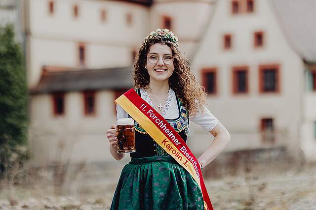 Die neue Forchheimer Bierkönigin Karolin I. in Tracht und der rot-gelben Schärpe in den Forchheimfarben. Auf der Schärpe steht: 11. Forchheim Bierkönigin Karolin I. In der rechten Hand hält sie einen Krug Bier. Im HIntergrund sieht man verschwommen die historische Kaiserpfalz.
