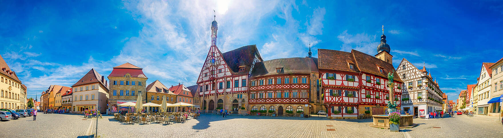 Rathausplatz Forchheim Panoramabild eines Altstadtplatzes mit Fachwerkensemble und Sandsteingebäuden bei strahlendem Sonnenschein. Links im Bild befindet sich eine Außengastronomie mit Sonnenschirmen und Bestuhlung. Rechts im Bild ein Brunnen.