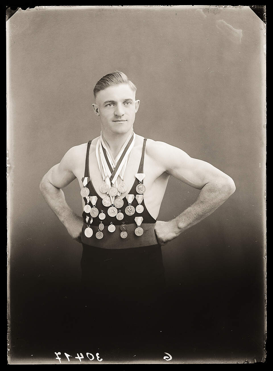 Eine schwarz-weiß Fotografie aus den 1920er Jahren zeigt einen erfolgreichen Ringer in Sportbekleidung, der seine vielen Medaillen präsentiert.
