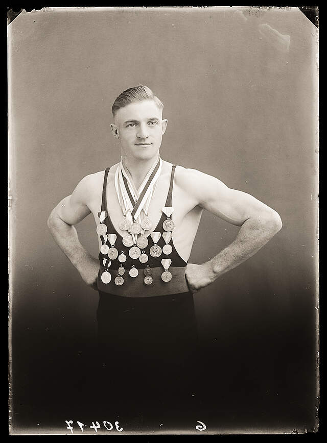 Eine schwarz-weiß Fotografie aus den 1920er Jahren zeigt einen erfolgreichen Ringer in Sportbekleidung, der seine vielen Medaillen präsentiert.