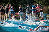 Schwimmer kraulen in einem Außenschwimmbecken. Am Beckenrand feuern Zuschauer die Sportler an.