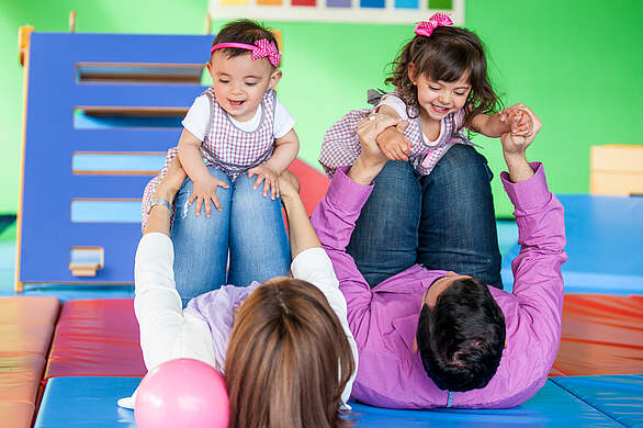 Zwei Eltern liegen mit angewinkelten Beinen auf Weichbodenmatten, während ihre beiden Kleinkinder auf ihren Schienbeinen liegen.