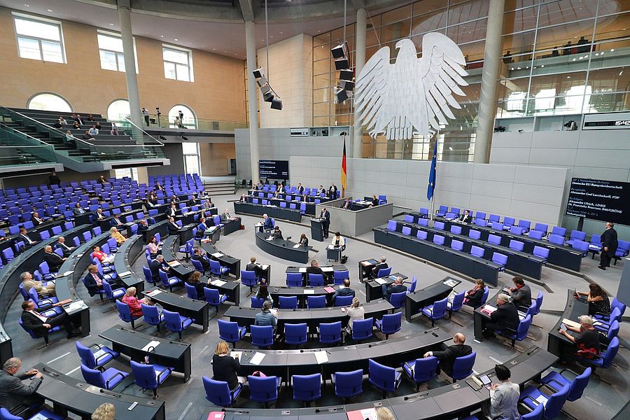 Blick in den Plenarsaal des Deutschen Bundestages mit im Halbkreis angeordneten blauen Stuhlreihen. Vorne oberhalb des Rednerpults befindet sich der Bundesadler.