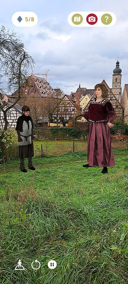 Screnshot aus der App Via Forte Screenshot einer Erlebnis-App, bei der zwei historische virtuelle Personen auf einer Wiese vor einer Altstadt stehen. Links im Bild eine Junge, rechts eine Frau in dunkelrotem Gewand.chnung in der linken Hand.