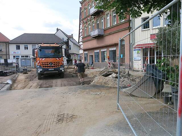 Ein Lkw steht auf einer Baustelle, bei der Pflasterflächen abgerochen wurden. Arbeiter und Bauzäune sowie Bau- und Bodenmaterialien sind zu sehen.
